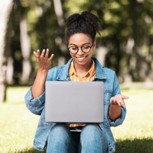 A young black woman with dark hair tied up wearing dark framed glasses, jeans, denim jacket and yellow and white shirt sitting on the grass with a laptop on her lap.