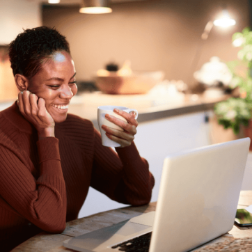 A black woman with short hair sits at a dining table smiling with her laptop open holding a white coffee mug.