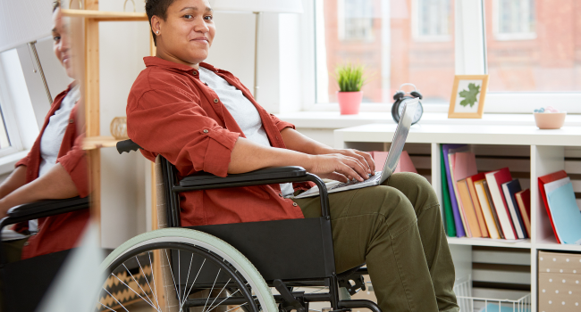 Black woman with short hair wearing a red shirt and green pants sitting in a wheelchair with a laptop on her lap.