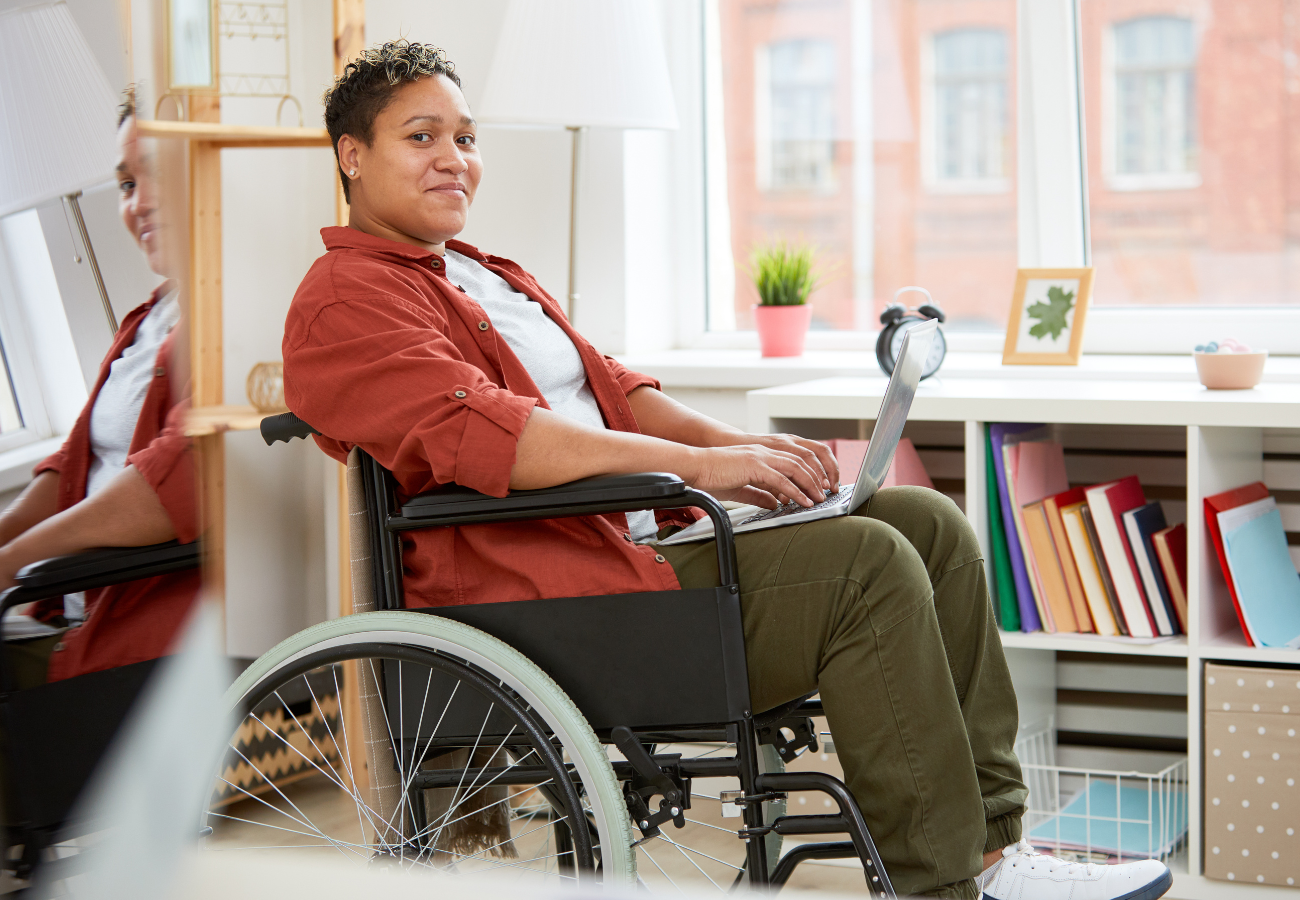 Black woman with short hair wearing a red shirt and green pants sitting in a wheelchair with a laptop on her lap.