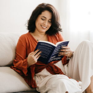 A white woman with thick dark shoulder length hair sits on a white lounge reading a book. She is wearing a burnt orange linen top with light linen pants.