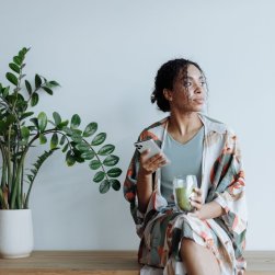 A woman of colour sits alone on a wooden bench next to an indoor plant. She has dark curly hair pulled back in a loose bun, is wearing a grey shirt and floral dressing gown and is holding a phone and glass of juice.