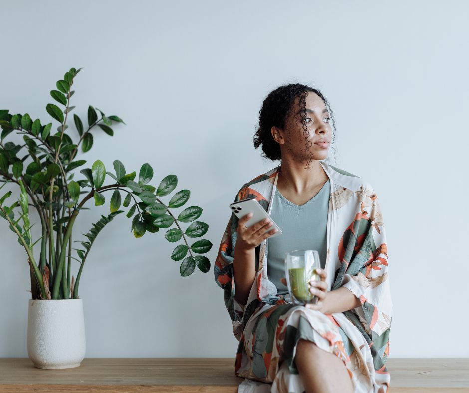 A woman of colour sits alone on a wooden bench next to an indoor plant. She has dark curly hair pulled back in a loose bun, is wearing a grey shirt and floral dressing gown and is holding a phone and glass of juice.