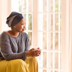 A South African woman sits in front of floor to ceiling windows. She is wearing a grey and ivory coloured head scarf, grey top and yellow skirt. She is holding a cup of coffee and gazing out the window.