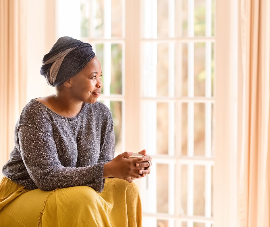 A South African woman sits in front of floor to ceiling windows. She is wearing a grey and ivory coloured head scarf, grey top and yellow skirt. She is holding a cup of coffee and gazing out the window.